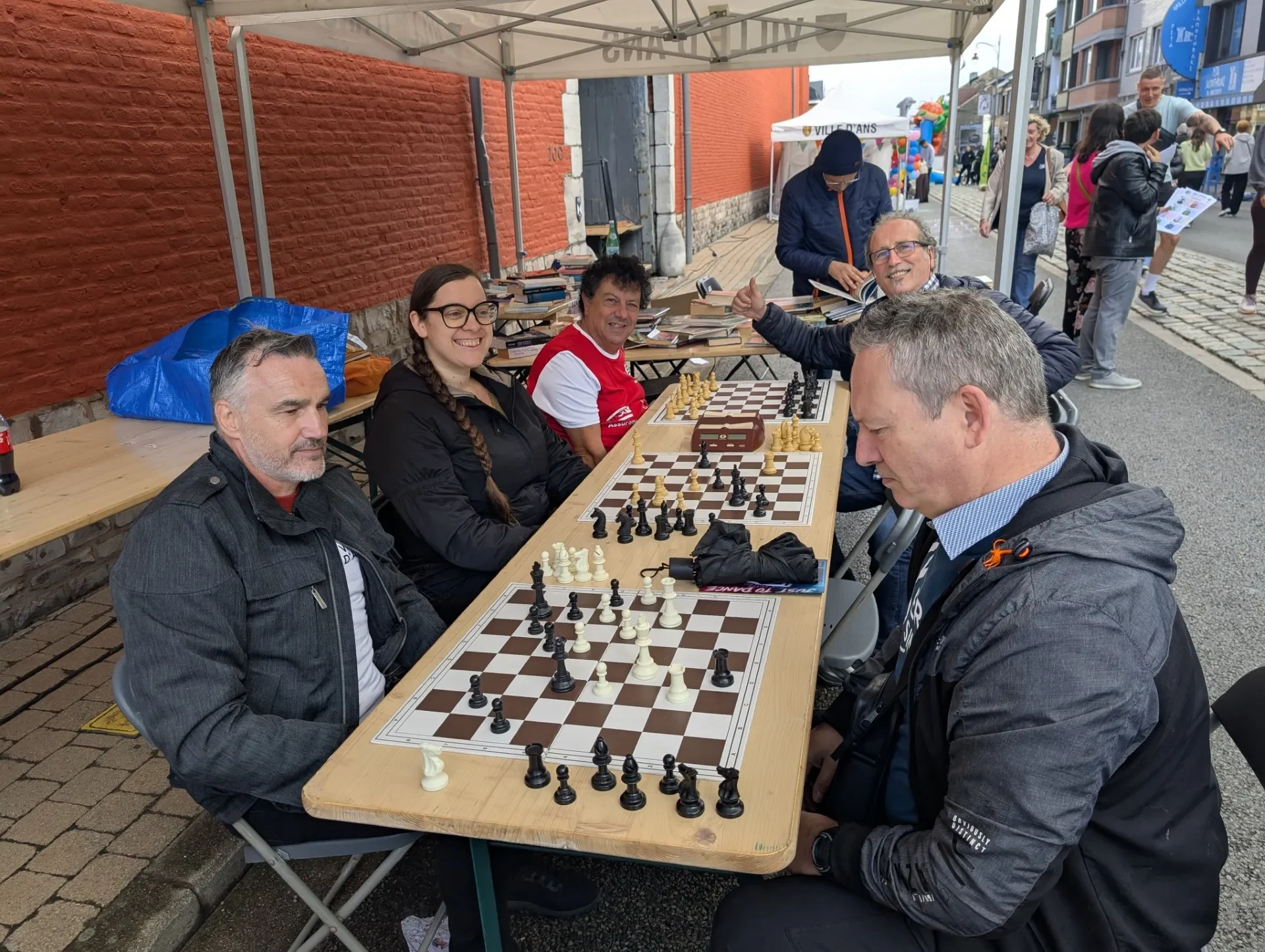 Quatre membres du club autour d'une longue table, deux d'entre eux jouant aux échecs avec une pendule, sous un chapiteau de la Ville d'Ans installé dans la rue ; un étal de livres et les passants de la journée sans voiture à l'arrière-plan.